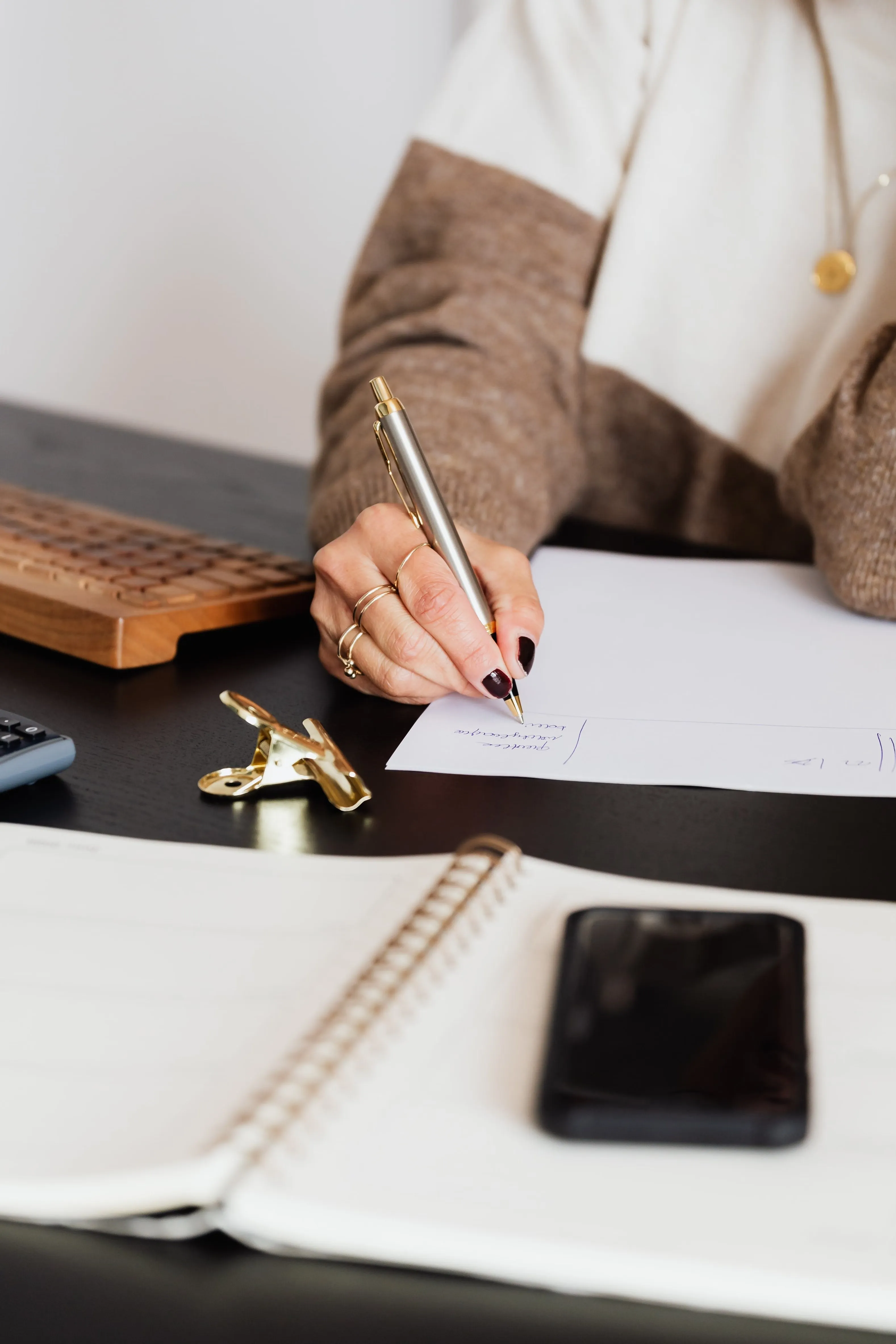 A hand writing notes with a gold pen at a desk, beside a wooden keyboard, a notebook and a phone.