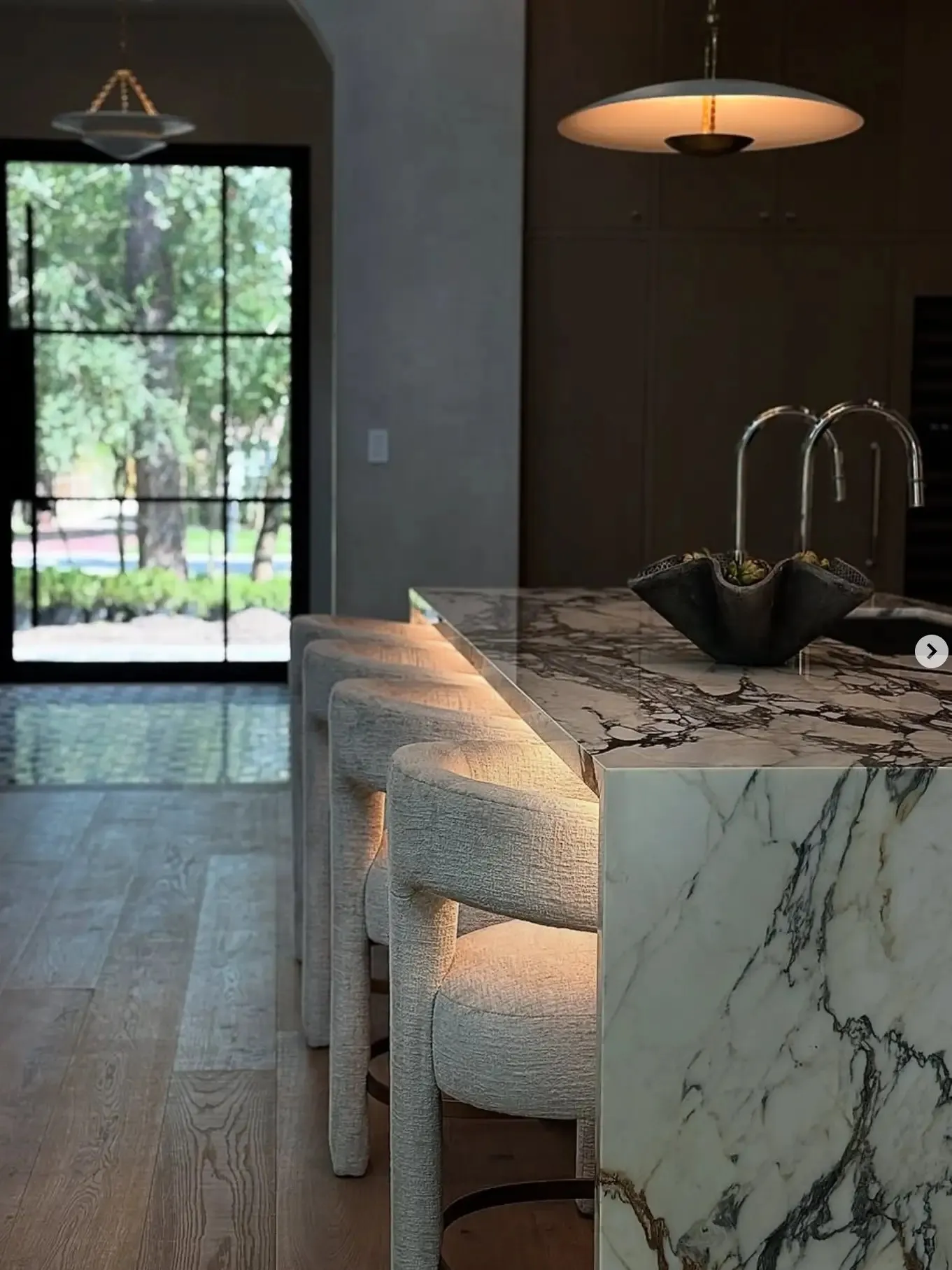 Luxe Design Group post: a moody kitchen with a veined-marble waterfall island, sculptural bouclé stools and a glowing pendant.