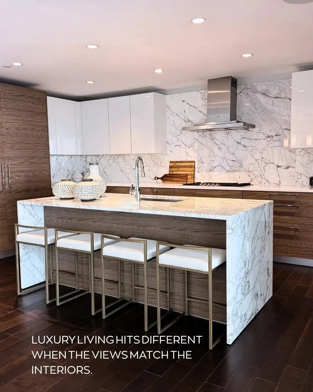 Luxe Design Group post: a white kitchen with a marble waterfall island, walnut cabinetry and four bar stools, captioned “Luxury living hits different when the views match the interiors.”